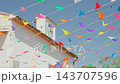 Church bell tower with colorful pennant flags fluttering across bright blue sky during sunny festival day, diagonal composition showing whitewashed building and terracotta roof tiles, lively outdoor,v 143707596