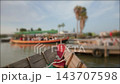 Weathered wooden boat bow with coiled rope and red cushion floating by busy marina with tour boat dock and palm trees in soft warm light, close foreground perspective, calm water reflections and dock 143707598