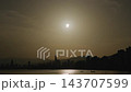 Partially eclipsed sun over silhouetted coastal city skyline with golden water reflections, distant mountains and small boats in calm hazy light, tranquil urban seascape at sunrise or dusk 143707599