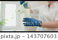 Gloved technician pouring viscous white liquid from measuring jug into clear beaker in bright clinical room, close midshot showing hands wristwatch and white uniform 143707603