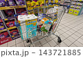 A shopping cart filled with everyday grocery items stands in a supermarket aisle. Rows of fully stocked store shelves form the background scene. 143721885