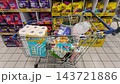 A shopping cart filled with everyday grocery items stands in a supermarket aisle. Rows of fully stocked store shelves form the background scene. 143721886