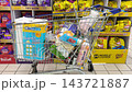 A shopping cart filled with everyday grocery items stands in a supermarket aisle. Rows of fully stocked store shelves form the background scene. 143721887