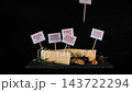 A male hand inserts E339, E234, E160b, E452, E407 and E202 markers into cheese beside walnuts, rosemary, grapes, bread and wine. Additive risk concept 143722294