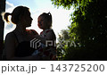 Beautiful silhouette of young mom kiss her cute small girl in summer garden. Happy parent and child sharing sweet emotional moment in nature. Motherhood, parental care and unconditional love concept 143725200