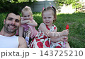 Portrait of happy young father with two little daughters smiling outdoor at sunny day. Cheerful family sit on green grass looking into camera in summer garden. Loving dad spend time with pretty kids 143725210