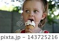 Portrait of attractive small girl eating delicious ice cream in a waffle cone outdoor. Beautiful child biting cold sweet dessert in summer garden. Cute kid enjoying tasty refreshing food on sunny day 143725216