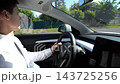 Profile of young businessman in formal wear driving modern electric car on urban road. Successful male entrepreneur commuting to work in a luxury electrical vehicle in city during a bright sunny day 143725256