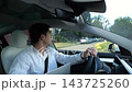Young manager driving modern electric car reversing out of his home driveway. Male entrepreneur keeps close eye on the mirrors and rearview camera ensuring safe start to morning commute to the office 143725260
