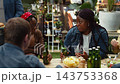 Friends pass plates to serve food and laugh around rooftop table, shared dining, casual hospitality and relaxed social connection outdoors. Cheerful group at BBQ event. 143753368