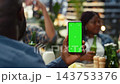 Vertical green screen mockup on mobile phone held by man at rooftop table as people socialize behind, combining replaceable screen with casual group setting. 143753376