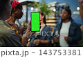 Vertical green screen on mobile phone held in front of friends at social gathering on rooftop, offering editable mobile display while guests mingle under warm decorative lights. 143753381