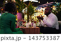 Girlfriend pays attention to her boyfriend telling stories during relaxed romantic evening. Close up of man and woman sharing red wine at rooftop dinner surrounded by lush foliage 143753387