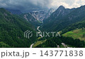 Wide drone shot of deep alpine valley surrounded by steep green forested slopes. Small isolated farmhouses dot valley floor under cloudy sky in Slovenia. 143771838