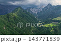 Wide drone shot of deep alpine valley surrounded by steep green forested slopes. Small isolated farmhouses dot valley floor under cloudy sky in Slovenia. 143771839