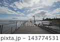 Wooden Boardwalk Leads Toward Distant Tower Over Tidal Flats And Calm Water, With Empty Rails And Broad Cloudy Sky In Peaceful Seaside Light, Following Shoreline 143774531