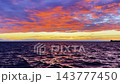 A vibrant crimson and orange sunset illuminates scattered storm clouds over dark waves, distant mountains, and a ship outline visible on the horizon. 143777450
