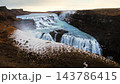 Gullfoss waterfall in Iceland with tiered cascades, mist rising from icy turquoise river, snow-dusted volcanic canyon walls, and warm evening light over dramatic remote wilderness landscape. 143786415