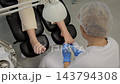Pedicurist tends to clients toenail with powered file during salon pedicure. Practitioner in blue gloves steadies foot as client sits back in professional treatment chair, filing and shaping toenail. 143794308