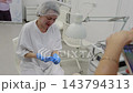 Beauty technician puts on blue gloves at pedicure workstation, smiling as she prepares for client care. Salon lamp, treatment tools and supplies stand beside seated technician in bright studio. 143794313
