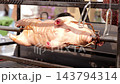 Whole hog rotates above grill during spit roasting, secured by metal forks and bar. Barbecue cooking turns pork over heat, showing browned skin and traditional rotisserie preparation. 143794314