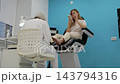 Woman receives pedicure as practitioner tends her bare foot in clinic chair. Client checks her smartphone during foot care, appearing relaxed in clean, brightly lit treatment room. 143794316