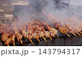 Vendor cooks dozens of meat skewers on charcoal grill, adjusting their position through billowing smoke. Busy street food preparation delivers sizzling, browned barbecue portions for serving. 143794317