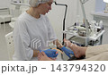 Pedicurist works on clients feet during salon pedicure, using small tools around toes while client rests in treatment chair. Foot care professional concentrates beneath bright task lighting. 143794320