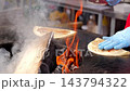 Cook tends flatbread over open fire on traditional griddle, pressing and turning dough as it cooks. Rising smoke and bright flames give this hands on bread making process energetic feel. 143794322