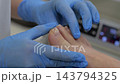 Close-up pedicure treatment in clinic with gloved technician inspecting glossy big toenail and cuticle area on client's foot under soft neutral lighting for hygienic nail care session 143794325