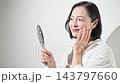 [White background] A Japanese woman in her 50s checks the skin on her cheeks in the mirror. Beautiful skin, skincare, anti-aging care, firmness and radiance. 143797660