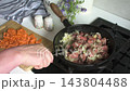 A woman stirs sliced beef liver in a pan while adding chopped carrots, onions, salt, and pepper. Fresh ingredients are visible on a cutting board nearby 143804488