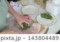 In a home kitchen during breakfast preparation, a woman arranges arugula, boiled eggs, and tomatoes on a plate beside a cutting board 143804489