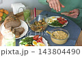 At home in the morning, a man serves stewed vegetables beside hummus, boiled eggs, arugula, tomatoes, bread, and a smoothie for breakfast 143804490