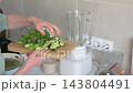 In a home kitchen at breakfast, a woman prepares a smoothie with cucumber, celery, chia seeds, and plant based milk using a blender 143804491