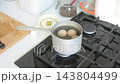 A hand lifts a saucepan with eggs above a gas burner in a home kitchen, with a wooden cutting board and jars on the counter 143804499