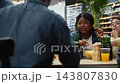 Woman holding beer bottle looks upward while friends talk around barbecue table on rooftop, capturing candid social moment, conversation and casual outdoor gathering. 143807830