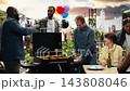Men smile and talk beside rooftop grill with corn and patties cooking, combining casual barbecue preparation, bottled drinks, conversation and outdoor hosting. 143808046