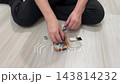 Hands assembling small electronic kit on floor, screwdriver and tiny screws placed beside clear case, person seated crosslegged, calm focused hobby repair scene with scattered components and toolkit 143814232