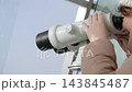 Tourist in winter clothing observes landmarks through binoculars from panoramic observation deck of tall building. Winter urban tourism during cold season. 143845487