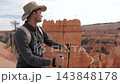 Caucasian male tourist wearing safari hat and backpack, leaning on trekking poles while admiring majestic red rock hoodoos at bryce canyon national park during sunny day 143848178