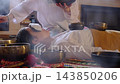 High angle view of a therapist playing a Tibetan singing bowl near a woman resting with an eye pillow on a colorful rug. Private sound bath rituals support deep relaxation, stress relief, and calm. 143850206