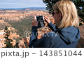 Blonde female traveler standing on cliff edge taking photos with smartphone at bryce canyon national park, capturing red rock hoodoo formations and scenic wilderness on sunny day 143851044