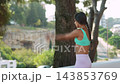 Outdoor fitness warmup: adult woman raises both arms overhead beside large tree on elevated urban terrace with bright natural light 143853769