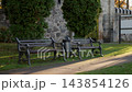 Empty park benches beside stone wall and sunlit walkway with evergreens and quiet garden setting behind them in late day 143854126