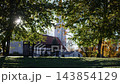 Empty park bench beneath sunlit autumn trees in quiet urban green space with grassy slope and blurred buildings behind 143854129