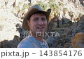 Cheerful male hiker in a cowboy hat filming a travel vlog while trekking through a rocky desert canyon, with wild palm trees, an oasis and sunny wilderness scenery 143854176