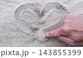 Close-up top view of man hand drawing by finger heart shape symbol on sand surface on sandy beach. Top-down first person POV of male hands. Summer vacation holiday and romantic love confession theme. 143855399