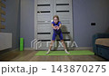 Young caucasian girl performs physical exercise on yoga mat indoors. Child engages in fitness activity during home workout, focused expression on face. Healthy lifestyle development. 143870275