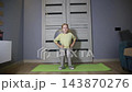 Young caucasian girl performing squats with exercise ball against wall, doing fitness workout on green yoga mat indoors at home, focusing on leg strength 143870276